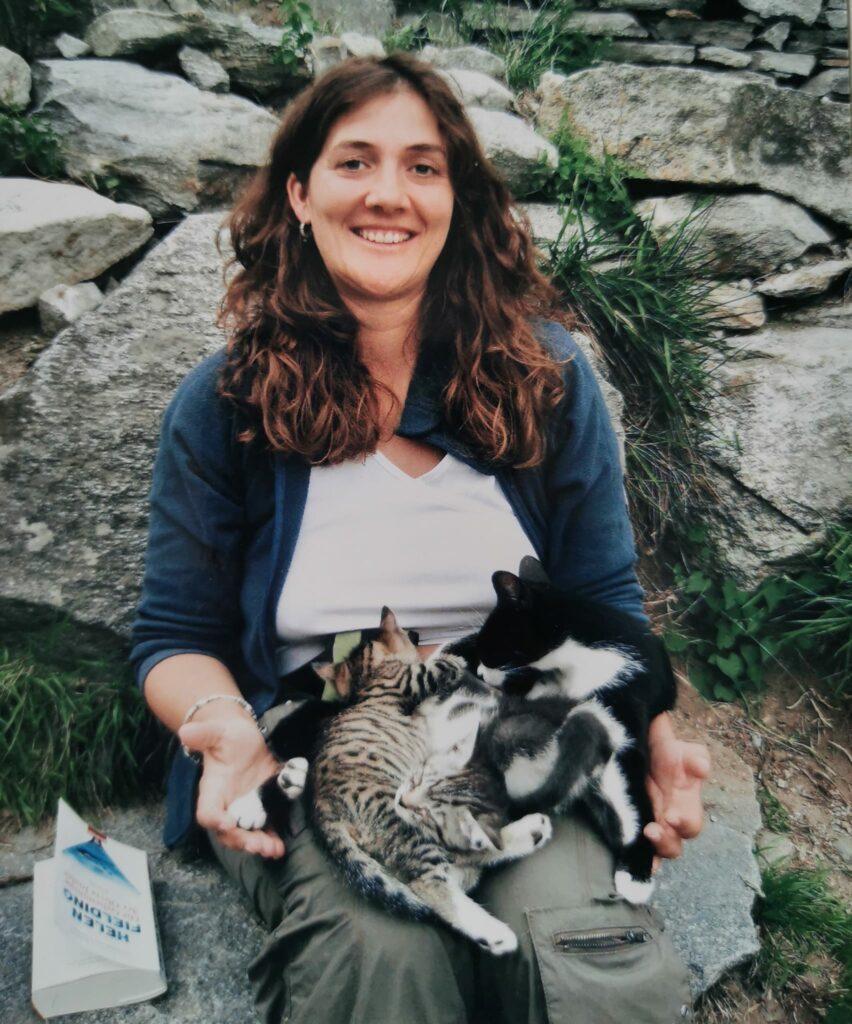 Frau mit langen braunen Locken sitzt mit einer Katze und ihren Jungen auf dem Schoß auf grauen Steinen 