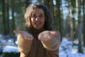 Eine Frau mit mittlelangen locken steht im Winterwald und streckt die Hände zum Betrachter aus. 